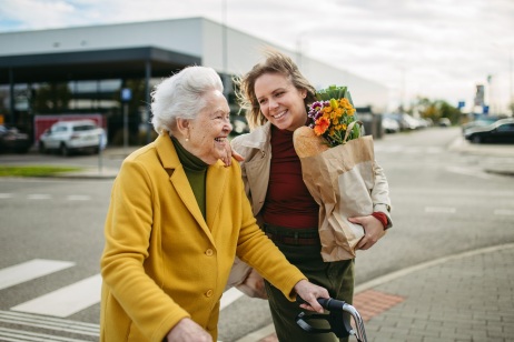 Eine junge Frau hält einen Blumenstraß und begleitet eine ältere Dame mit Rollator über die Straße. 