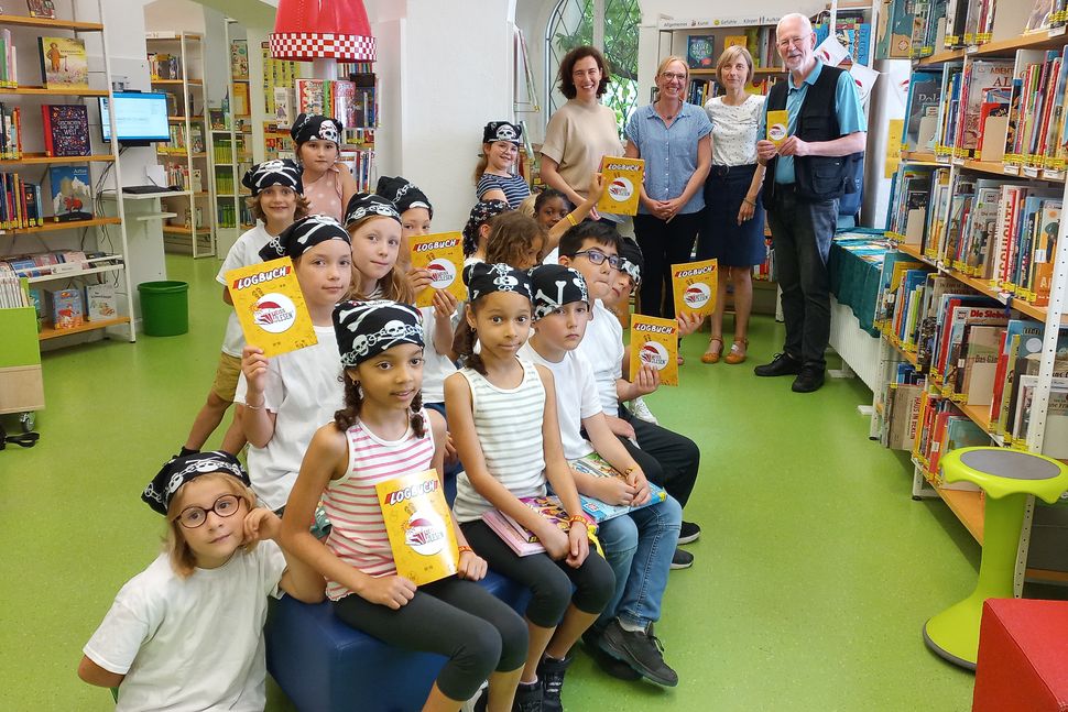 Schulklasse einer Grundschule in einem Bibliotheksraum mit Erwachsenen im Hintergrund. Die Schüler tragen Piratenkopftücher und haben ein gelbes Logbuch in der Hand.